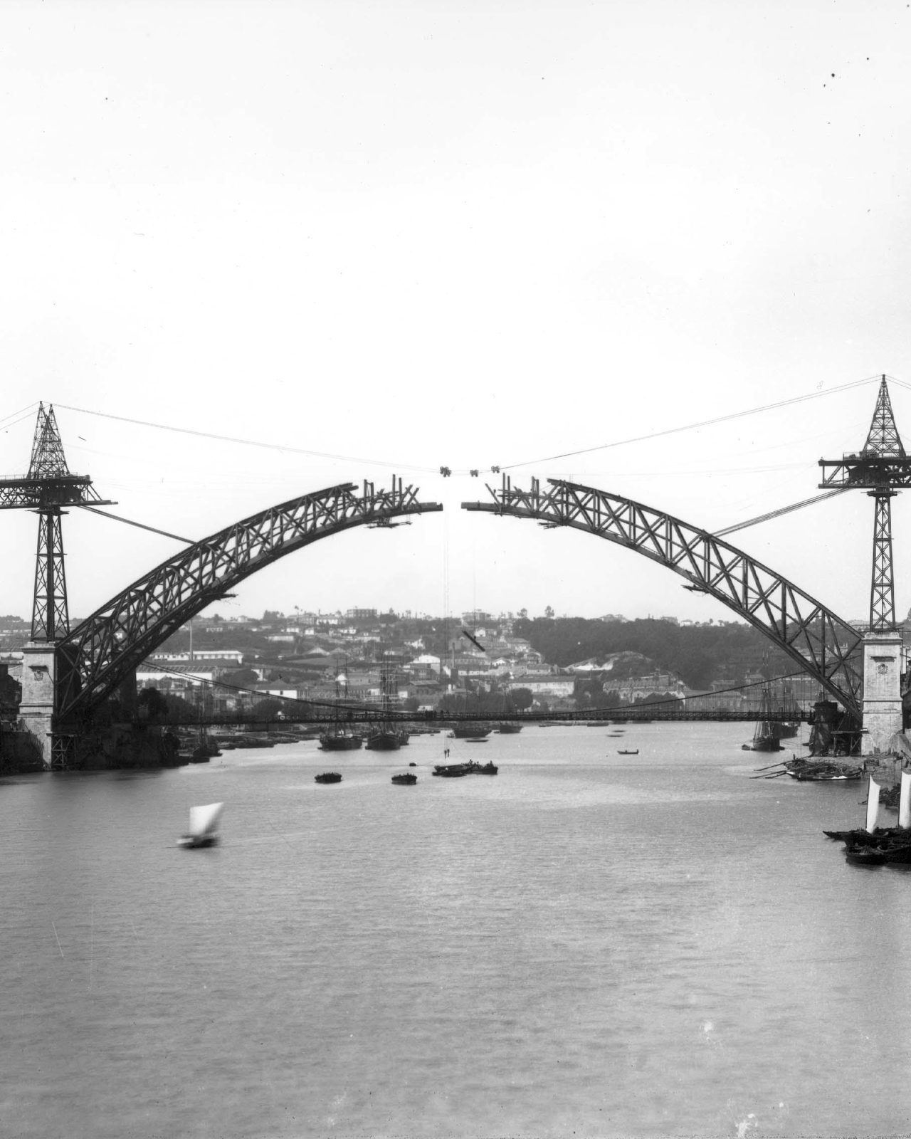 Fotografia de Emílio Biel, 1883. As duas metades do arco da ponte Luiz I, penduradas dos cabos das duas torres de montagem erguidas sobre os pilares, ainda sem se tocarem ao centro. Por baixo, a velha ponte pênsil continua a trabalhar, e há barcos no rio.